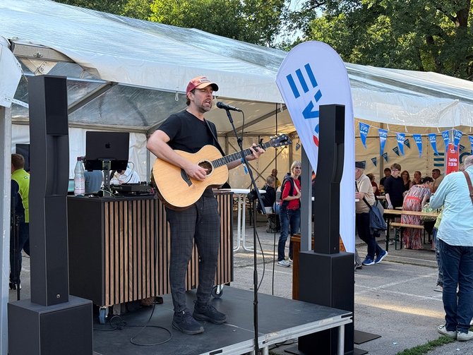 Eine Person steht auf einer kleinen Bühne und spielt Gitarre, während sie in ein Mikrofon singt. Im Vordergrund steht eine Person auf einer erhöhten Plattform und hält eine akustische Gitarre. Die Person singt in ein Mikrofon, das an einem Ständer befestigt ist. Auf beiden Seiten der Bühne stehen große, schwarze Lautsprecher. Hinter der Person befindet sich ein Tisch mit Technik, darunter ein Laptop und ein Mischpult. Rechts neben der Bühne steht eine hohe, schmale Fahne mit blauem und rotem Aufdruck. Im Vordergrund rechts stehen mehrere Tische mit Gegenständen wie Kartons, verpackten Produkten und Textilien. Im Hintergrund ist ein großes, weißes Zelt aufgebaut. Unter dem Zelt sitzen und stehen mehrere Menschen an Tischen und Bänken. Es sind außerdem weitere Fahnen und Wimpel mit Schriftzügen zu sehen. Hinter dem Zelt stehen hohe Bäume bei sonnigem Wetter.