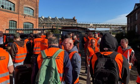 Gruppe von mehreren Dutzend Personen in orangefarbenen Warnwesten auf einem gepflasterten Hof zwischen Backsteingebäuden. Einige Personen tragen Rucksäcke oder Kappen. Im Hintergrund sind Industriegebäude, ein überdachter Bereich und eine erhöhte Stahlkonstruktion zu sehen. Sonniger Himmel.