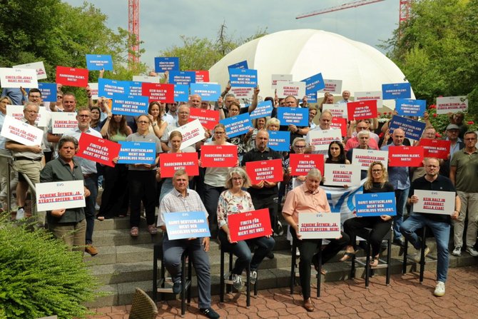 Große Gruppe von Menschen steht im Freien und hält vor sich oder über den Kopf zahlreiche rechteckige Schilder in den Farben Blau, Rot und Weiß. Auf den Schildern sind kurze Slogans in deutscher Sprache zu lesen, jeweils mit „EVG“ als Absender. Einige Personen halten zusätzlich eine blaue Fahne mit weißer Beschriftung. Im Hintergrund sind Pflanzen, ein Gebäude mit heller Fassade und gestapelte Holzscheite zu sehen. Die Personen sind unterschiedlich gekleidet, stehen dicht beieinander und blicken überwiegend zur Kamera.