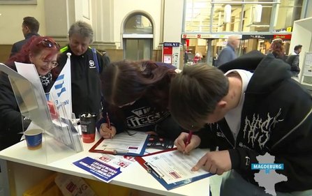 In einer Bahnhofshalle stehen mehrere Personen an einem kleinen Informationsstand. Zwei Personen beugen sich über Klemmbretter und füllen Formulare mit Stiften aus. Auf dem Tisch liegen weitere Formulare, Flyer und ein Becher. Hinter dem Stand stehen weitere Personen; mehrere Gesichter sind verpixelt. Im Hintergrund sind Bahnhofselemente wie Wände, Durchgänge und Beschilderung sichtbar. Unten rechts ist der Schriftzug „MAGDEBURG“ eingeblendet.