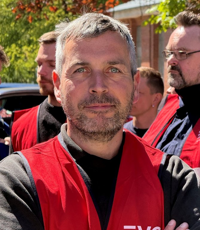 Nahaufnahme einer Person im Vordergrund mit roter Warnweste und dunklem Oberteil, die Arme vor dem Körper verschränkt. Im Hintergrund stehen weitere Personen in roten Warnwesten und blauer Arbeitskleidung. Dahinter sind ein Baum mit grünem und orangefarbenem Laub sowie ein Backsteingebäude erkennbar.