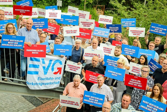 Große Gruppe von Menschen steht im Freien vor Bäumen und hält zahlreiche Schilder in die Kamera. Die Schilder sind überwiegend rot, blau und weiß und tragen kurze Texte sowie das „EVG“-Logo. Einige Personen heben die Schilder hoch, andere halten sie auf Brusthöhe. Ein großes Banner mit „EVG Eisenbahn- und Verkehrsgewerkschaft“ ist im Vordergrund zu sehen. Die Gruppe steht dicht beieinander auf einer gepflasterten Fläche mit Geländer im Vordergrund und dichter Vegetation im Hintergrund.