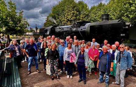Gruppe von etwa 35 Personen auf einem gepflasterten Platz vor einer schwarzen Dampflokomotive. Die Lokomotive steht seitlich im Hintergrund zwischen hohen Bäumen, aus einem Bereich oberhalb des Kessels steigt etwas Dampf oder Rauch auf. Links und rechts sind Bänke und weitere Bahnanlagen sichtbar. Über der Szene hängen dunkle Wolken.