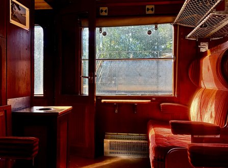Abteil eines historischen Eisenbahnwagens mit dunkler Holzverkleidung. Rechts stehen rot gestreifte Polstersessel, darüber ein Gepäcknetz. In der Mitte befindet sich ein großes Fenster mit Blick auf Bäume und eine helle Außenfläche. Links sind eine Tür, eine kleine Tischfläche und ein gerahmtes Foto an der Wand zu sehen.