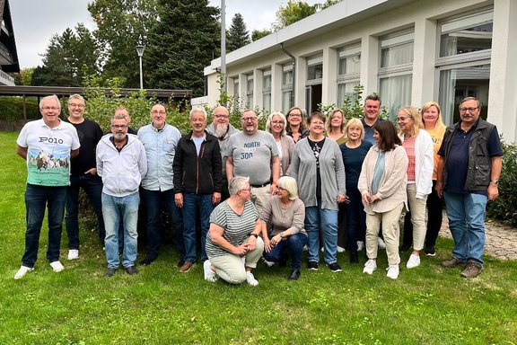 Gruppenfoto von etwa 20 Personen auf einer Rasenfläche vor einem Gebäude mit großen Fenstern. Die Personen sind in zwei Reihen für ein gemeinsames Foto aufgestellt. Die meisten stehen nebeneinander, zwei Personen befinden sich im Vordergrund in kniender Position. Die Gruppe trägt überwiegend Freizeitkleidung wie Jeans, Pullover, Jacken und Hemden. Im Hintergrund stehen Büsche und kleinere Bäume vor einem hellen Gebäude mit großen Fensterflächen und weißen Vorhängen.