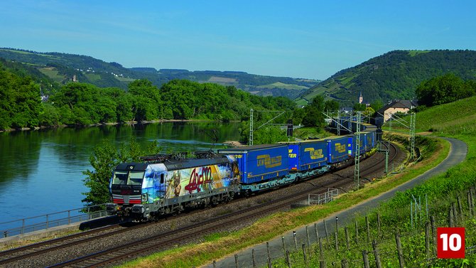 Elektrolokomotive mit großflächiger Werbebeklebung zieht mehrere blaue Containerwagen auf einer zweigleisigen Bahnstrecke entlang eines Flusses. Die Strecke verläuft durch eine grüne Hügellandschaft mit Bäumen, Weinbergen und einem parallel verlaufenden Weg. Unten rechts befindet sich eine rote Markierung mit der Zahl „10“.
