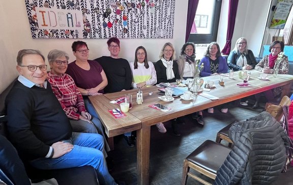 Eine Gruppe von zehn Personen sitzt dicht nebeneinander an einem langen Holztisch in einem Café oder Restaurant. Auf dem Tisch stehen mehrere leere und halbvolle Tassen, Gläser, Kannen, Besteck sowie gefaltete Servietten und einige Flyer. Hinter der Gruppe hängt ein großes, gemustertes Wandbild mit einzelnen bunten Dekorationselementen. Durch zwei Fenster im Hintergrund fällt Tageslicht in den Raum.