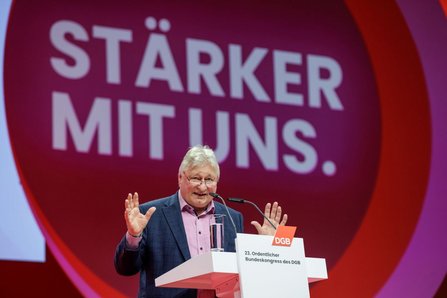 Person steht hinter einem Rednerpult und hebt beide Hände. Auf dem Pult befinden sich ein Mikrofon und ein Glas Wasser. Auf der Vorderseite des Pults ist ein rotes Logo mit der Aufschrift „DGB“ sowie der Text „23. Ordentlicher Bundeskongress des DGB“ zu sehen. Im Hintergrund ist eine große runde Fläche in Rot- und Violetttönen mit dem weißen Schriftzug „STÄRKER MIT UNS.“ zu erkennen.