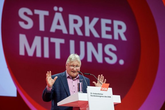 Person steht hinter einem Rednerpult und hebt beide Hände. Auf dem Pult befinden sich ein Mikrofon und ein Glas Wasser. Auf der Vorderseite des Pults ist ein rotes Logo mit der Aufschrift „DGB“ sowie der Text „23. Ordentlicher Bundeskongress des DGB“ zu sehen. Im Hintergrund ist eine große runde Fläche in Rot- und Violetttönen mit dem weißen Schriftzug „STÄRKER MIT UNS.“ zu erkennen.