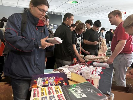 Innenraum mit einem Informationsstand. Im Vordergrund liegt ein Tisch mit Broschüren, Flyern, Schlüsselbändern, gefalteten weißen Textilien mit rotem Aufdruck und einem Becher mit Deckel und Strohhalm. Mehrere Personen stehen rund um den Tisch und betrachten oder halten die ausgelegten Materialien. Im Hintergrund sind weitere Personen und Automaten an der Wand zu sehen.