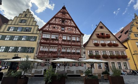 Mehrere historische Gebäude mit steilen Giebeln an einem Platz. Das mittlere Fachwerkhaus trägt die Beschriftungen „Hotel Deutsches Haus“ und „Altdeutsches Restaurant“. Vor den Gebäuden stehen Sonnenschirme, Pflanzkübel und Sitzbereiche. Darüber sind blauer Himmel und Wolken zu sehen.