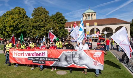 Demonstrationszug auf einer Grünfläche vor einem Gebäude mit Arkadengang und Kuppeldach. Mehrere Personen tragen Fahnen und ein großes rotes Banner mit der Aufschrift „Hart verdient! Deine Arbeit. Deine Gesundheit. Deine Rente.“. Auf dem Banner sind zwei aneinanderstoßende Fäuste sowie die Logos „DGB“ und „MACH DICH STARK MIT UNS“ zu sehen. Im Hintergrund stehen weitere Fahnen, Bäume und ein rotes Fahrzeug. Der Himmel ist blau mit dünnen Wolkenstreifen.