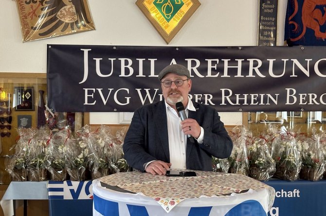 Eine einzelne Person steht hinter einem runden Tisch und hält ein Handmikrofon. Auf dem Tisch liegt eine gemusterte Tischdecke, darunter ist ein blau‑weißes Tuch mit sichtbarem „EVG“-Schriftzug angebracht. Im Hintergrund hängt ein dunkles Banner mit der Aufschrift „Jubilarehrung EVG Wupper Rhein Berg“. Dahinter stehen mehrere in Folie verpackte Blumen- oder Geschenkarrangements auf Tischen. An den Wänden sind Wappen und Fahnen angebracht; der Raum ist hell beleuchtet.
