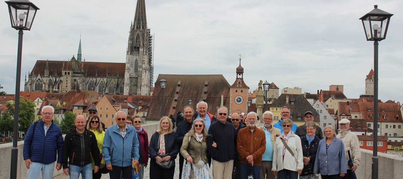 Eine Gruppe von etwa 18 Personen steht auf einer Brücke vor einer Altstadt mit vielen alten Häusern und einem großen Dom im Hintergrund. Im Vordergrund stehen mehrere Personen dicht nebeneinander auf einer breiten Steinbrücke. Einige tragen Jacken in Blau, Grau, Braun oder Beige, manche halten Taschen oder kleine Gegenstände in den Händen. Hinter der Gruppe verläuft die Brücke weiter nach hinten, mit hellen Steinplatten und Geländern an den Seiten. Auf beiden Seiten der Brücke stehen hohe Straßenlaternen. Im Hintergrund ist eine historische Stadt zu sehen: viele Häuser mit roten und braunen Dächern, unterschiedlich hoch und dicht gebaut. Besonders auffällig ist ein sehr großer, gotischer Dom mit zwei hohen Türmen und vielen Details an der Fassade. Daneben steht ein Gebäude mit einem Turm und einer großen Uhr. Der Himmel ist bedeckt, das Licht wirkt gleichmäßig und eher kühl.