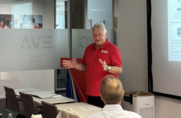Eine Person in einem roten Poloshirt steht vor einer Projektionsfläche und spricht zu sitzenden Teilnehmenden. Neben der Projektionsfläche stehen ein Rednerpult und ein Luftreinigungsgerät. Hinter der Person befinden sich Glaswände mit mattierten Bereichen und teilweise sichtbaren Schriftzügen. Auf der Projektionsfläche sind Text und Abbildungen zu erkennen.