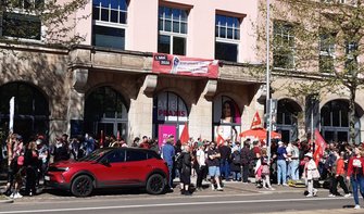 Straßenszene vor einem mehrstöckigen Gebäude mit Arkaden. Vor dem Gebäude steht eine größere Menschenmenge, einige halten Fahnen. Über den Arkaden hängt ein rotes Banner mit der Aufschrift „1. Mai 2026 – Erst unsere Jobs, dann eure Profite“. Auf der Straße davor ist ein rotes Auto geparkt, Bäume werfen Schatten auf Fassade und Gehweg.
