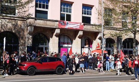 Straßenszene vor einem mehrstöckigen Gebäude mit Arkaden. Vor dem Gebäude steht eine größere Menschenmenge, einige halten Fahnen. Über den Arkaden hängt ein rotes Banner mit der Aufschrift „1. Mai 2026 – Erst unsere Jobs, dann eure Profite“. Auf der Straße davor ist ein rotes Auto geparkt, Bäume werfen Schatten auf Fassade und Gehweg.