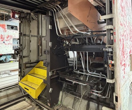 Blick auf das Innere einer technischen Anlage mit Metallrahmen, Leitungen, Kabeln und einem schwarzen Gehäuse. Links befinden sich gelbe Metallstufen, im Hintergrund sind Schienen und weitere Maschinenteile sichtbar.
