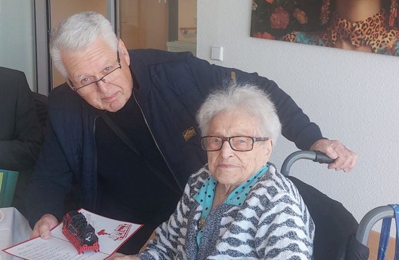 In einem Innenraum stehen zwei Personen dicht beieinander. Eine Person sitzt auf einem Stuhl mit sichtbaren Armlehnen, die andere steht dahinter und hält eine Hand an die Rückenlehne. Die stehende Person trägt eine dunkle Jacke und hält in der linken Hand ein Blatt Papier mit roter Umrandung und einem kleinen roten Zug darauf. Die sitzende Person trägt eine gemusterte Jacke über einem hellen Oberteil. Im Hintergrund sind eine helle Wand, eine Tür mit Glasfläche und ein gerahmtes Bild mit farbigem Motiv zu sehen.