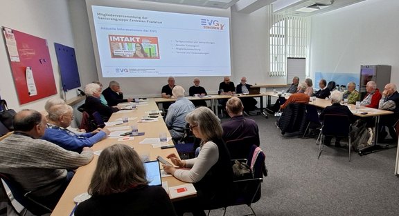 Großer, heller Seminarraum mit U-förmig angeordneten Tischen. An den Tischen sitzen zahlreiche Personen. Auf den Tischen liegen Papierunterlagen, Notizblöcke, Stifte, Gläser und Trinkflaschen. An der Stirnseite steht eine Person hinter einem Tisch mit mehreren weiteren sitzenden Personen. An der Wand ist eine projizierte Präsentationsfolie zu sehen mit lesbarem Text: „Mitgliederversammlung der Seniorengruppe Zentrale Frankfurt“, „Aktuelle Informationen der EVG“ sowie Logos und der Schriftzug „EVG Senioren“. An den Wänden hängen farbige Pinnwände, rechts ist ein großes Fenster mit senkrechten Lamellen.