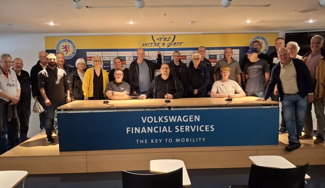 Eine Gruppe von etwa 20 Personen steht und sitzt hinter einem langen Tisch mit drei Mikrofonen in einem Konferenzraum. Auf der Vorderseite des Tisches steht „Volkswagen Financial Services – The Key to Mobility“. Im Hintergrund befindet sich eine Wand mit Sponsorenlogos und einem Schriftzug „Herz unserer Stadt“ sowie zwei runde Vereinslogos.