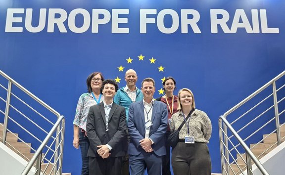 Gruppenfoto mit sechs Personen auf einer Treppe vor einer großen blauen Wand. Oben auf der Wand steht in großen weißen Buchstaben „EUROPE FOR RAIL“. Hinter der Gruppe ist ein Kreis aus gelben Sternen zu sehen. Die Personen tragen überwiegend Business- oder Business-Casual-Kleidung sowie Namensschilder. Rechts und links verlaufen Metallgeländer entlang der Treppen. Die Aufnahme ist frontal und symmetrisch ausgerichtet.