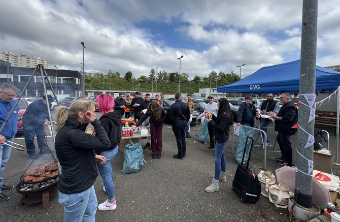 Außenbereich eines Parkplatzes mit mehreren stehenden Personen rund um einen Tisch mit Getränkekisten und Flaschen. Links ein runder Grill mit Grillgut und aufsteigendem Rauch. Rechts ein blaues Faltzelt mit der Aufschrift „EVG“. Mehrere geparkte Autos im Hintergrund, Metallzäune und Gebäude. Der Boden ist asphaltiert, der Himmel ist bewölkt.