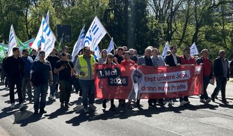 Eine weite Aufnahme einer Menschenmenge auf einer Straße, umgeben von Bäumen. Viele Personen tragen und schwenken weiße Fahnen mit blauen Streifen und der Aufschrift „EVG“. In der Mitte der Gruppe wird erneut das rote Banner mit der Aufschrift „1. Mai 2026“ und „Erst unsere Jobs, dann eure Profite“ getragen. Die Straße, Gehwege und eine grüne Parkumgebung sind sichtbar, das Tageslicht wirft klare Schatten auf den Boden.