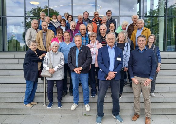 Gruppenfoto von etwa 30–35 Personen, die auf mehreren Stufen einer Treppe vor einem Gebäude mit großer Glasfassade stehen. Die Personen sind in mehreren Reihen angeordnet, tragen Alltagskleidung in verschiedenen Farben und Mustern und blicken zur Kamera. Im Vordergrund stehen mehrere Personen mit verschränkten oder nebeneinanderliegenden Händen; im Hintergrund sind weitere Personen dicht nebeneinander auf den Stufen. Hinter der Gruppe spiegeln die großen Fensterscheiben Bäume und den bewölkten Himmel; im Innenbereich sind runde Deckenleuchten sichtbar. Die Treppe besteht aus hellgrauen Steinplatten mit klaren Kanten.