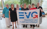 Acht Personen stehen in einem hellen Innenraum vor großen Fensterflächen und halten gemeinsam ein weißes Banner. Auf dem Banner steht in großen blauen und roten Buchstaben „EVG“ sowie darunter „Eisenbahn- und Verkehrsgewerkschaft“. Im Hintergrund sind Pflanzen, Sitzgelegenheiten und weitere teilweise sichtbare Personen zu sehen. Die Personen tragen überwiegend dunkle oder farbige Oberteile und stehen in einer Reihe hinter dem Banner.
