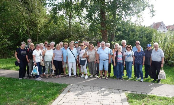 Gruppe von etwa 25 Personen steht auf einem gepflasterten Weg im Freien. Im Vordergrund befinden sich Rasenflächen und ein Weg aus rechteckigen Pflastersteinen. Hinter der Gruppe stehen Bäume und dichtes Grün. Am rechten Bildrand ist teilweise ein Fachwerkgebäude sichtbar.