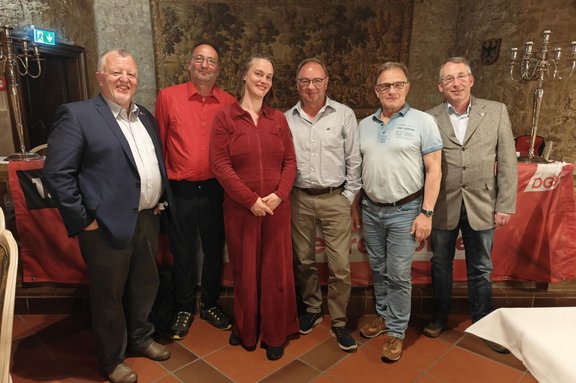 Sieben Personen stehen nebeneinander in einem historischen Innenraum mit Steinwänden und gemustertem Wandteppich. Alle tragen unterschiedliche Kleidung, darunter Sakkos, Hemden, Polohemd und ein einfarbiger Overall; die Farben reichen von Blau, Grau und Beige bis Rot. Die Personen stehen auf einem Fliesenboden in Terrakotta-Farben. Hinter der Gruppe ist ein rotes Banner mit weißer Aufschrift „DGB“ teilweise sichtbar. Links an der Wand befindet sich ein beleuchtetes grünes Notausgangsschild. Rechts und links stehen hohe Kerzenleuchter. Ein weiß gedeckter Tisch ist am rechten Bildrand angeschnitten zu sehen.