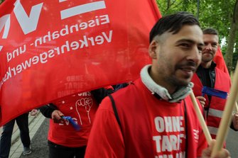Nahaufnahme eines Demonstrationszuges. Im Vordergrund trägt eine Person ein rotes T‑Shirt mit weißem Schriftzug, daneben sind rote EVG-Fahnen zu sehen. Weitere Personen mit roten T‑Shirts, Warnwesten und blauen Tröten befinden sich im Hintergrund entlang eines Weges mit Bäumen.