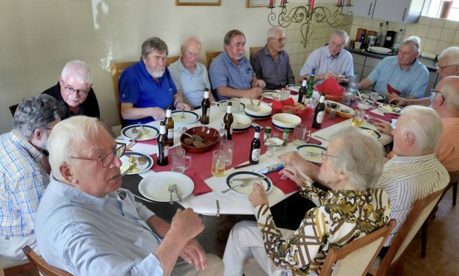 Mehrere Personen sitzen an einem Esstisch während einer Mahlzeit. Auf dem Tisch stehen Schüsseln, Teller, Gläser, Bierflaschen und Servietten. Der Tisch ist mit einer weißen Decke und einem roten Läufer gedeckt. Im Hintergrund sind eine Küchenzeile und weitere Sitzplätze zu sehen.