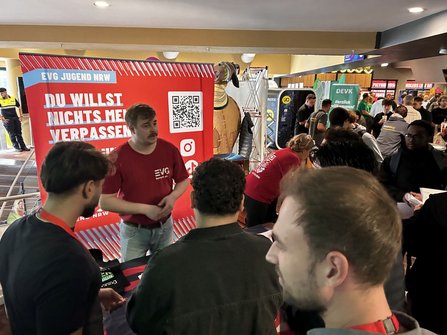 Innenraum mit Messeständen und vielen Personen. Im Vordergrund steht ein roter Aufsteller mit der Aufschrift „EVG-JUGEND NRW“ und „DU WILLST NICHTS MEHR VERPASSEN“ sowie einem QR-Code. Mehrere Personen befinden sich an einem Informationsstand mit Tisch und Werbematerial. Im Hintergrund sind weitere Stände, Banner und Automaten sichtbar.