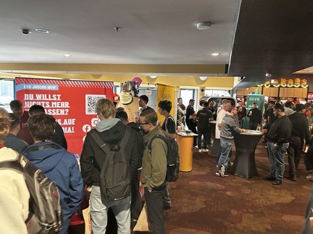 Innenraum mit mehreren Informationsständen und zahlreichen Personen. Links steht ein großer roter Aufsteller mit der Aufschrift „EVG JUGEND NRW“, einem QR-Code und teilweise sichtbarem Text „DU WILLST NICHTS MEHR VERPASSEN?“. In der Mitte und rechts stehen Messe- und Beratungsstände, darunter Stände mit den Beschriftungen „BAHN BKK“ und „DEVK“. Mehrere Stehtische sind im Raum verteilt. Viele Personen tragen Rucksäcke oder Umhängetaschen. Die Beleuchtung ist warm, der Boden dunkel gemustert.
