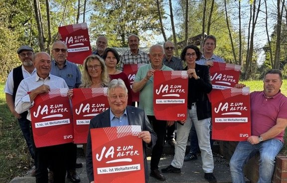 Gruppe von etwa 13 Personen steht und sitzt im Freien auf einem Weg zwischen Bäumen. Mehrere Personen halten rote Plakate mit der Aufschrift „JA ZUM ALTER“ und darunter „1.10. ist Weltaltentag“. Unten auf den Plakaten ist das Logo „EVG“ sichtbar. Im Vordergrund sitzt eine Person mit einem Plakat, weitere Personen stehen dahinter mit weiteren Plakaten. Im Hintergrund sind Bäume, Grünflächen und ein kleines Gebäude zu sehen.