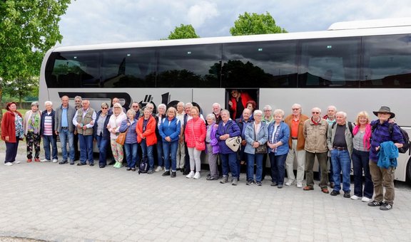 Große Gruppe von etwa 30 Personen steht in einer Reihe vor einem weißen Reisebus auf einem gepflasterten Platz im Freien. Der Bus füllt den Hintergrund fast vollständig aus, mit dunklen getönten Fenstern und sichtbarem Schriftzug auf der Seite. Die Personen tragen unterschiedliche Jacken, Westen und Freizeitkleidung in verschiedenen Farben und stehen dicht nebeneinander. Einige halten Taschen oder Rucksäcke in den Händen. Eine Person steht in der geöffneten Bustür. Im Hintergrund sind grüne Bäume und ein bewölkter Himmel zu sehen.