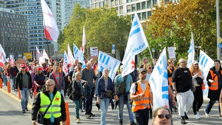 Großer Demonstrationszug auf einer Straße mit vielen weiß-blauen Fahnen und einigen roten Fahnen. Mehrere Personen tragen Warnwesten. Entlang der Straße stehen Bäume, Verkehrszeichen und moderne Gebäude mit Glas- und Wohnfassaden.