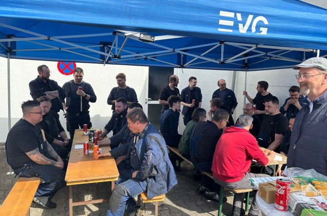 Mehrere Personen sitzen und stehen unter einem blauen Faltzelt mit der Aufschrift „EVG“. Vor dem Zelt stehen Holzbänke und Tische mit Getränkeflaschen, Dosen und Lebensmitteln. Auf einem Tisch liegen Servietten und Verpackungen. Im Hintergrund eine helle Wand mit einem runden Verkehrszeichen. Der Bereich wirkt wie ein improvisierter Aufenthalts- oder Pausenplatz im Freien.