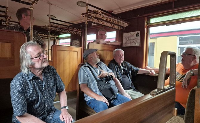 Innenraum eines historischen Wagenabteils mit Holzverkleidung, Holzbänken und Gepäckablagen aus Netzgeflecht. Sechs Personen sitzen oder stehen im Abteil. Durch die Fenster ist ein weiteres Fahrzeug in Gelb und Rot zu sehen. An der Wand hängt ein kleiner Netzplan.