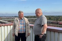 Zwei Personen stehen auf einer Brücke vor einem Geländer und unterhalten sich. Im Hintergrund erstreckt sich ein weitläufiges Bahngelände mit vielen Gleisen, Güterwagen, Lokomotiven und einer Brücke. Darüber befindet sich ein überwiegend klarer Himmel.