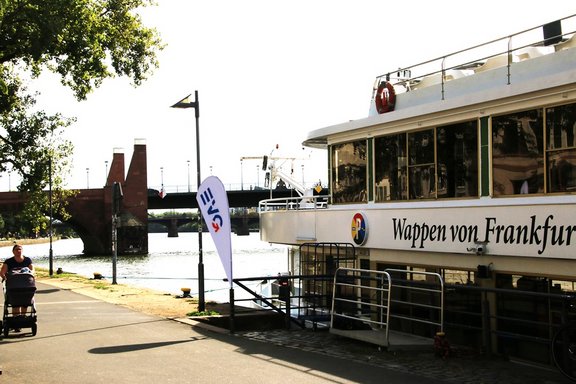 Ein Fahrgastschiff mit der Aufschrift „Wappen von Frankfurt“ liegt an einem Anleger am Flussufer. Daneben verläuft ein Weg, im Hintergrund sind eine Brücke und weitere Uferbauten zu sehen. Das Schiff nimmt den rechten Bildteil ein und ist mit großen Fenstern sowie einem Rettungsring auf dem Oberdeck ausgestattet. Vor dem Anleger steht eine weiße Fahne mit blau-rotem Logo. Links verläuft ein asphaltierter Uferweg entlang des Wassers. Auf dem Weg befindet sich eine Person mit einem Kinderwagen oder Rollwagen. Mehrere Bäume spenden Schatten am Bildrand.