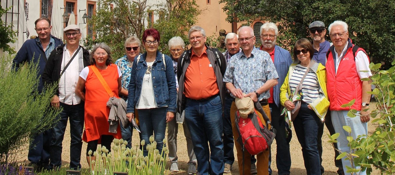 Eine Gruppe von etwa zwölf Personen steht in einem Garten vor einem Gebäude und blickt in Richtung Kamera. Die Personen stehen dicht nebeneinander auf einem sandigen Weg zwischen Pflanzen. Im Vordergrund sind blühende Gartenpflanzen und Kräuter zu sehen. Mehrere Personen tragen Jacken, Hemden oder Westen in verschiedenen Farben, darunter Rot, Orange, Blau und Gelb. Einige haben Rucksäcke, Taschen oder Kopfbedeckungen dabei. Auf der rechten Seite ist eine rote Weste mit der Aufschrift „EVG“ erkennbar.