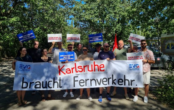 Gruppe von etwa 13 Personen steht im Freien vor Bäumen und hält ein großes weißes Banner. Auf dem Banner steht in großen Buchstaben: „Karlsruhe braucht Fernverkehr.“ Links und rechts auf dem Banner ist das EVG-Logo zu sehen. Mehrere Personen halten Schilder mit den Aufschriften „DAMIT DEUTSCHLAND NICHT DEN ANSCHLUSS VERLIERT.“ sowie „SCHIENE ÖFFNEN: JA. STÄDTE OPFERN: NEIN!“. Eine rote Fahne ist hinter der Gruppe sichtbar. Sonniger Tag mit Schatten auf dem Boden, im Hintergrund Bäume und teilweise parkende Fahrzeuge.