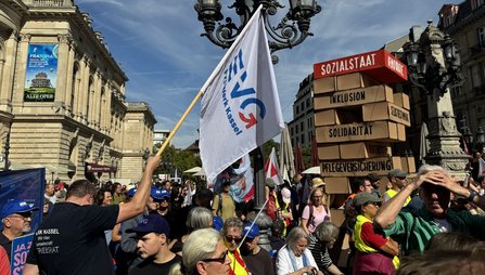 Menschenmenge auf einem Platz neben einem historischen Gebäude. Im Vordergrund wird eine weiße EVG-Fahne gehalten. Rechts steht ein großes Modell aus übergroßen Bausteinen mit den sichtbaren Aufschriften „SOZIALSTAAT“, „INKLUSION“, „SOLIDARITÄT“ und „PFLEGEVERSICHERUNG“. Mehrere Fahnen und Schilder sind über die Menge verteilt.