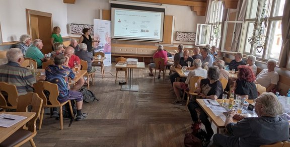 Ein Raum mit Holzboden, mehreren Tischen und Stühlen, an denen zahlreiche ältere Menschen sitzen. Vorne steht eine Person neben einer Leinwand mit projizierter Präsentation. Neben der Leinwand steht ein Roll-Up-Banner. Die Teilnehmenden schauen zur Präsentation oder unterhalten sich. An den Tischen stehen Gläser und Unterlagen. Große Fenster an der rechten Seite lassen Tageslicht in den Raum.