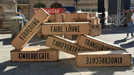 Auf einem gepflasterten Platz sind große quaderförmige Kartonattrappen wie umgestürzte Bausteine angeordnet. Sichtbare Aufschriften auf den Kartons sind „FAIRE LÖHNE“, „FÖRDERUNG“, „TARIFVERTRÄGE“, „TRANSFERSYSTEM“, „KINDERRECHTE“ und „VERTEILUNG“. Im Hintergrund stehen weitere Kartonattrappen, Absperrgitter, eine Bühne sowie Gebäude mit Steinfassaden.