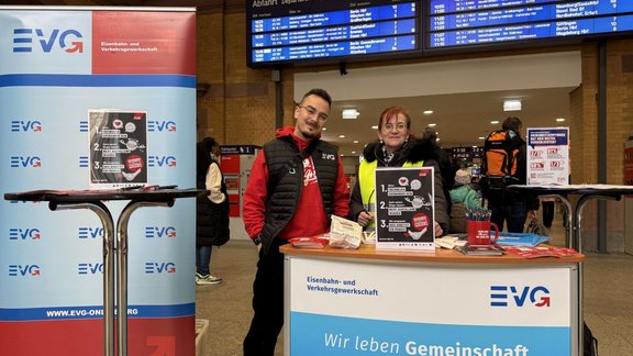 Informationsstand der Eisenbahn- und Verkehrsgewerkschaft (EVG) in einer Bahnhofshalle: Zwei Personen stehen hinter einem Tisch mit EVG-Logo, darauf liegen Flyer, Broschüren und Werbematerial. Daneben ein Roll-up-Banner und ein Stehtisch mit weiteren Infomaterialien; im Hintergrund sind eine Abfahrtstafel, Reisende und Bahnhofsarchitektur zu sehen.