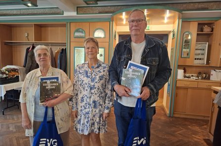 Drei Personen stehen nebeneinander in einem holzvertäfelten Raum mit türkisfarbenen Zierleisten. Die linke und die rechte Person halten jeweils ein Buch mit dem Titel „Eisenbahnen im Harz“, eine blaue Glückwunschkarte und eine blaue Tasche mit weißem EVG-Logo. Im Hintergrund sind eine Garderobe mit Jacken, ein Tisch mit Blumenstrauß, Schränke, Spiegel, eine kleine Küchenzeile und ein beleuchteter Durchgang zu sehen.